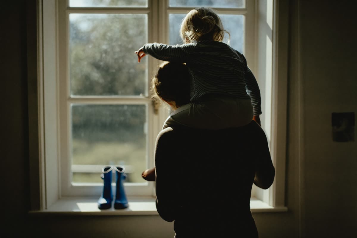 Elternteil mit Kind auf den Schultern schaut aus dem Fenster
